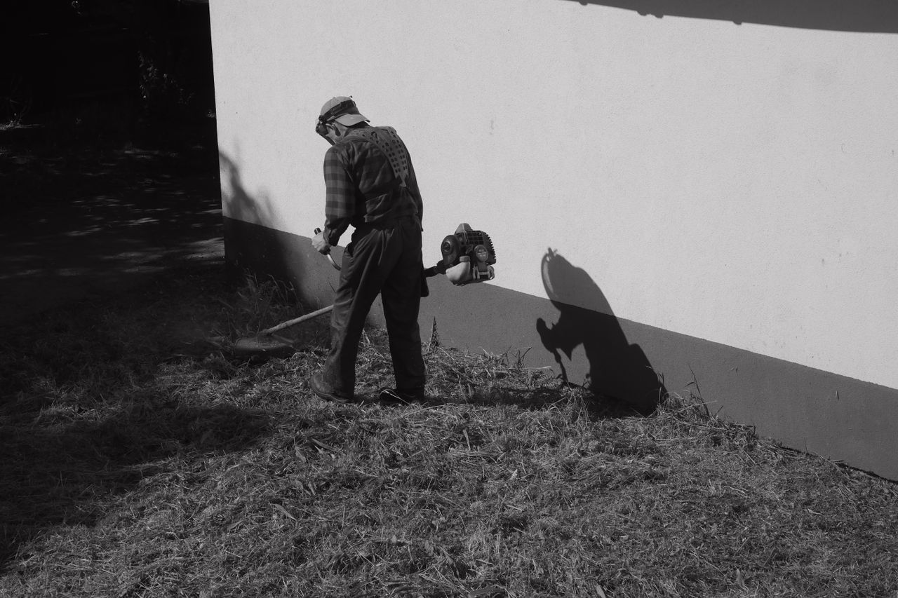 gallery-2 Black and white photo of a person trimming grass with a weed whacker next to a wall.