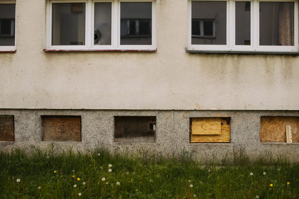 Exterior view of an abandoned building with boarded windows and overgrown grass.